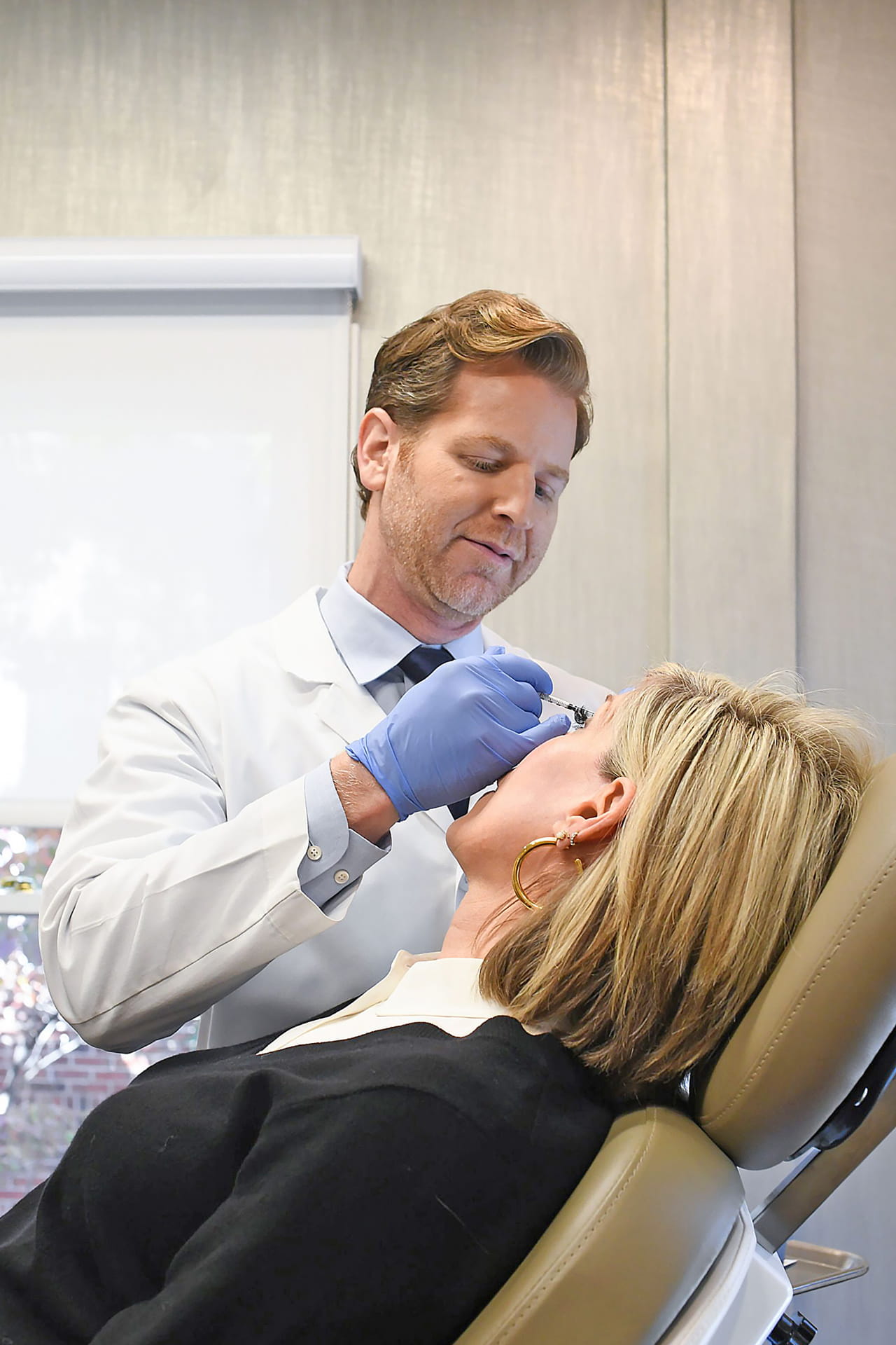 A doctor in a white coat and blue gloves performs a procedure on a patient in a clinical setting. The patient, with blonde hair, is reclining in a chair and has her eyes closed. The doctor is holding an instrument close to the patient's face, appearing to focus intently on the task. The background features a window with natural light filtering in.