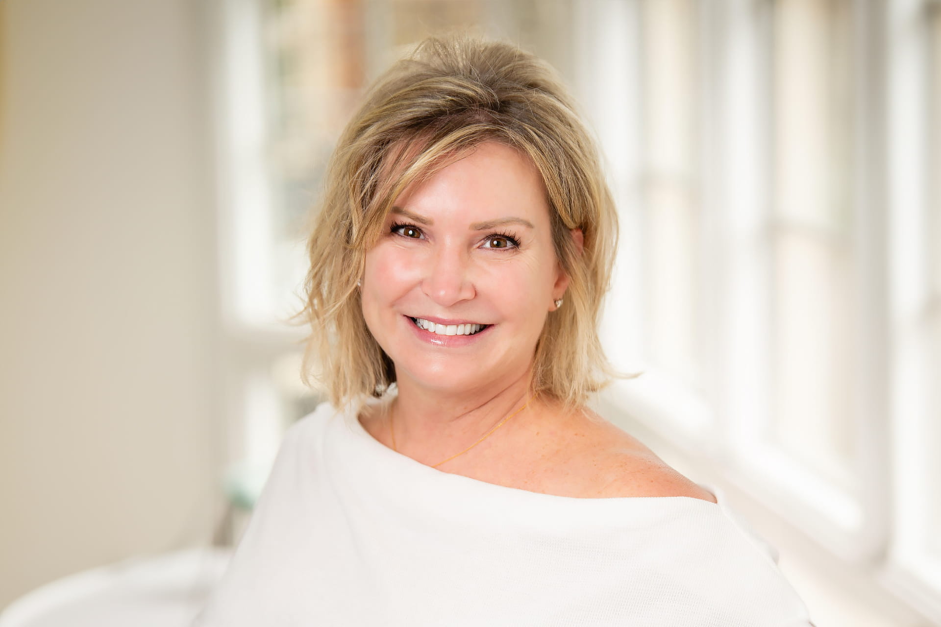 A woman with short blonde hair and a white off-shoulder top smiles at the camera. She is standing in a bright room with large windows in the background, allowing natural light to illuminate the space.