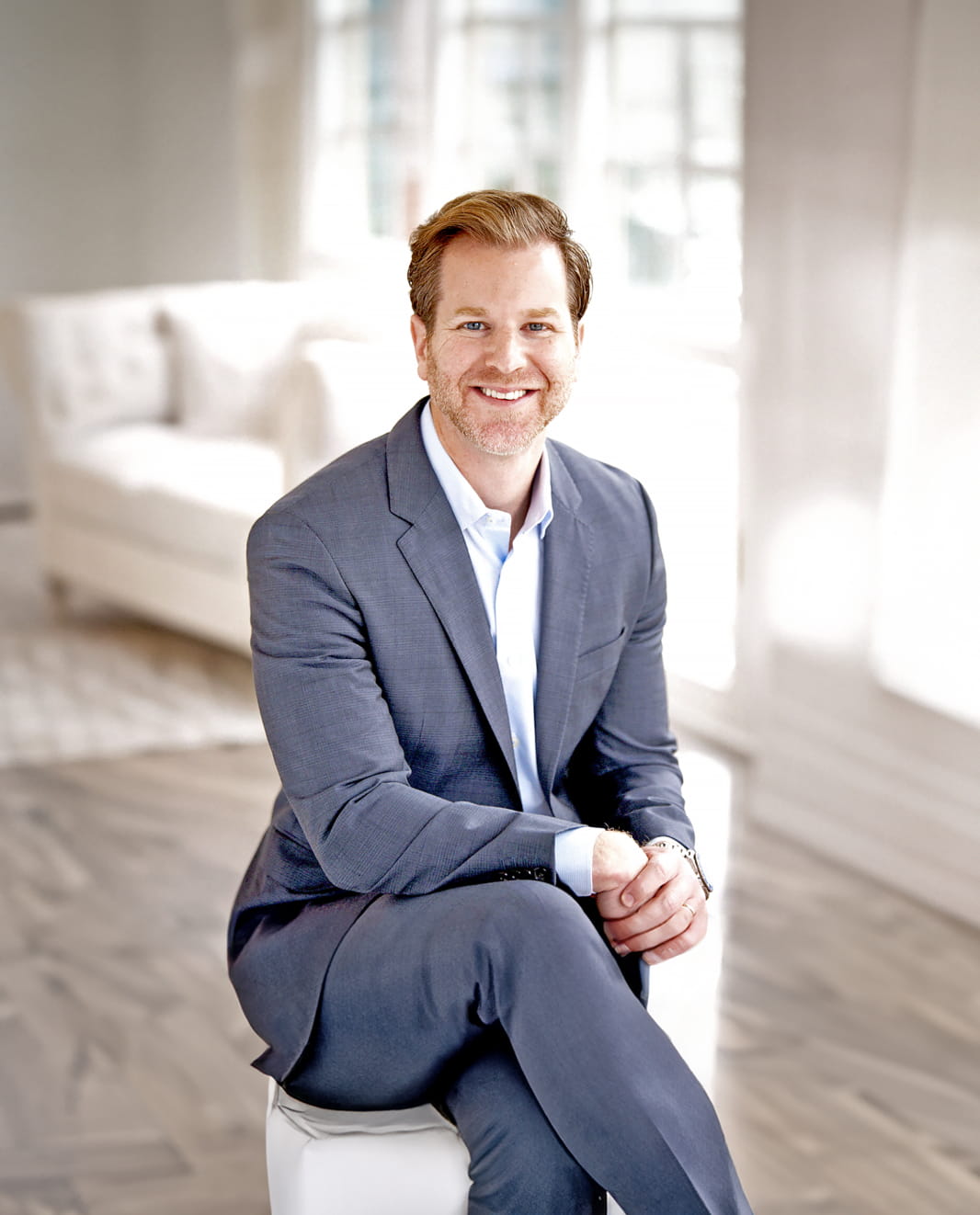 A man in a grey suit is sitting on a stool in a brightly lit room with a modern white interior. He is smiling and has his hands folded on his lap. There is a white couch and large windows in the background.