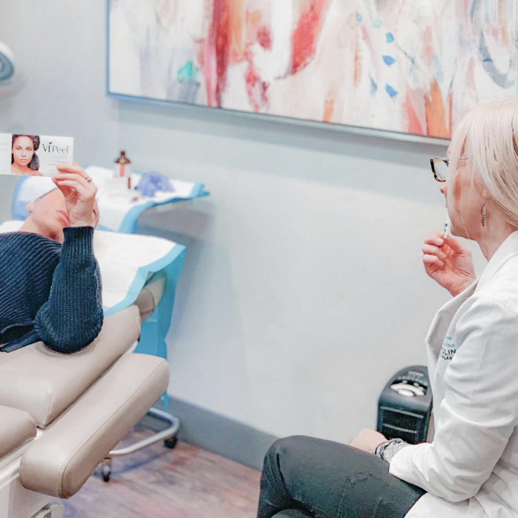 A person is lying on a treatment chair holding a pamphlet in front of their face. A medical professional wearing a white coat is seated next to them, discussing something. The room has modern decor with an abstract painting on the wall and medical equipment around.