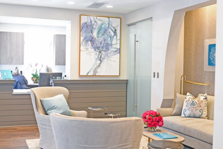 A modern dental office waiting room featuring two armchairs and a cushioned bench with decorative pillows. The space is decorated with a large abstract painting, a glass door, and a vase of pink flowers on the coffee table. A reception desk with two staff members working on computers is visible in the background. The room has a clean, contemporary design with soft neutral colors and wood flooring.