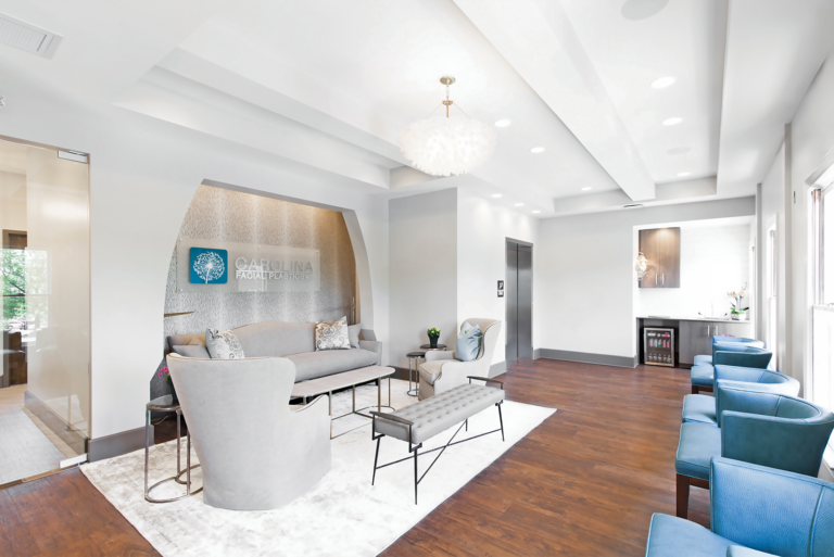 A modern and bright waiting room with a blend of contemporary and classic furniture. There are gray armchairs, a gray sofa, and a cushioned bench arranged around a coffee table on a large white rug. On the wall behind the sofa is a logo with the text "CAROLINA" and "PROJECT HEALTH" along with a tree graphic. The room has wooden flooring, blue chairs along one wall, and a small beverage station with a mini-fridge near the corner. Large windows allow natural light