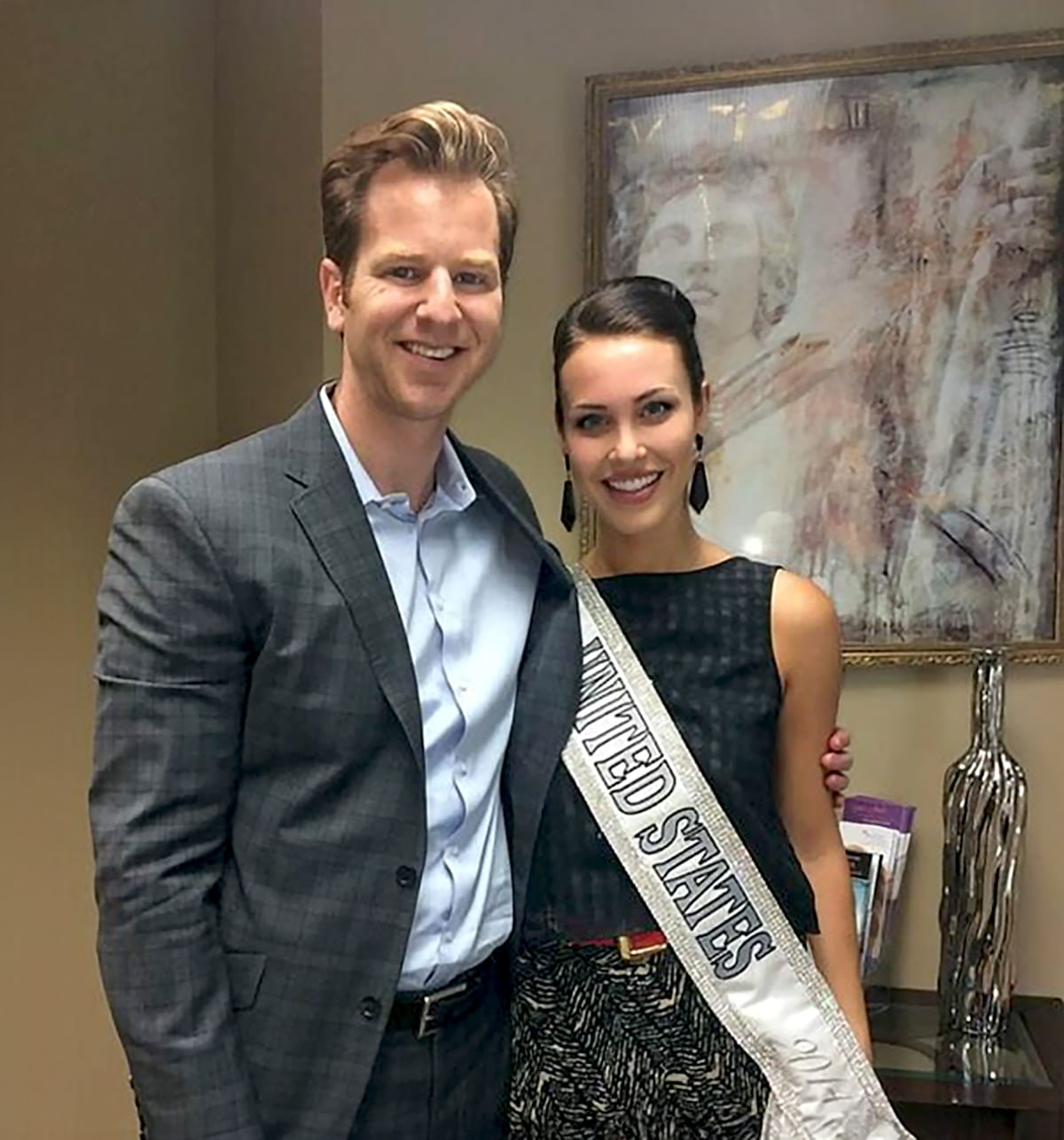 A man and a woman stand side by side indoors. The man, wearing a gray checkered blazer and light blue shirt, has his arm around the woman. The woman is wearing a dark sleeveless top and a sash that reads "United States." Both are smiling at the camera, and there is an abstract painting and a decorative object in the background.