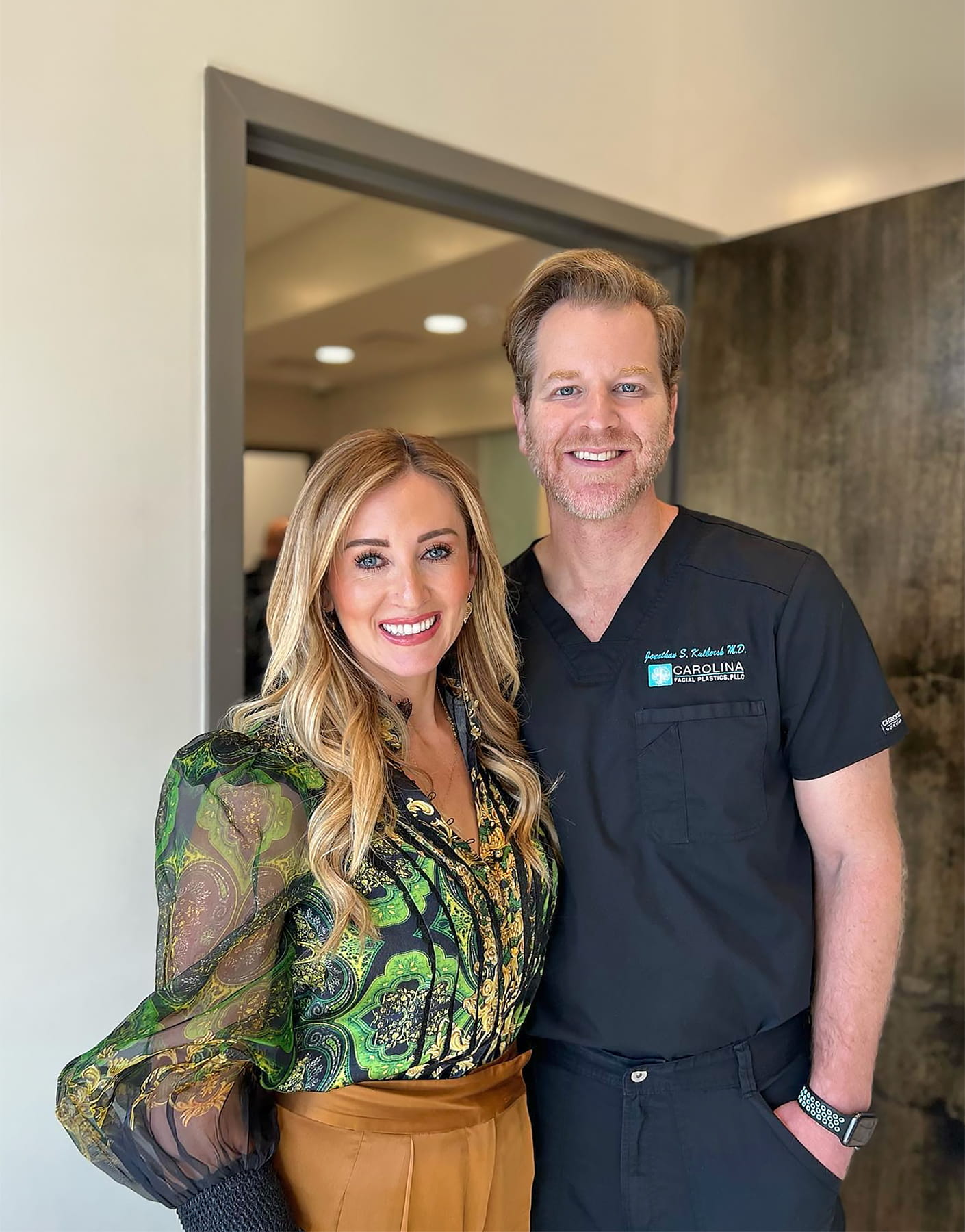 A woman with long blonde hair, wearing a green patterned blouse and mustard-colored pants, stands next to a man with short light brown hair in black medical scrubs. They are smiling and posing together in an indoor setting near a doorway.