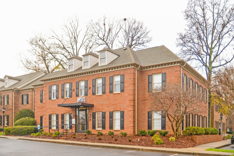 A two-story brick office building with a central entrance, black shutters, and windows evenly spaced across its facade. The front of the building features a small covered porch with stairs leading up to the entrance. The surrounding landscape includes manicured bushes and trees, with some of the trees bare of leaves, indicating a likely autumn or winter setting. A paved parking lot is visible in the foreground.