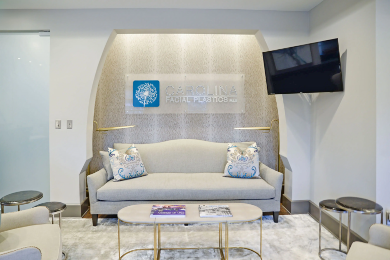 A modern waiting area features a light gray sofa adorned with patterned blue and white throw pillows. In front of the sofa, there are two contemporary-style coffee tables displaying magazines. The backdrop includes an illuminated logo reading "Carolina Facial Plastics" mounted on a textured wall panel. A flat-screen TV is mounted on the wall to the right, while a clear glass door is seen on the left. The space is decorated with neutral tones and elegant lighting fixtures, creating a sophisticated ambiance.