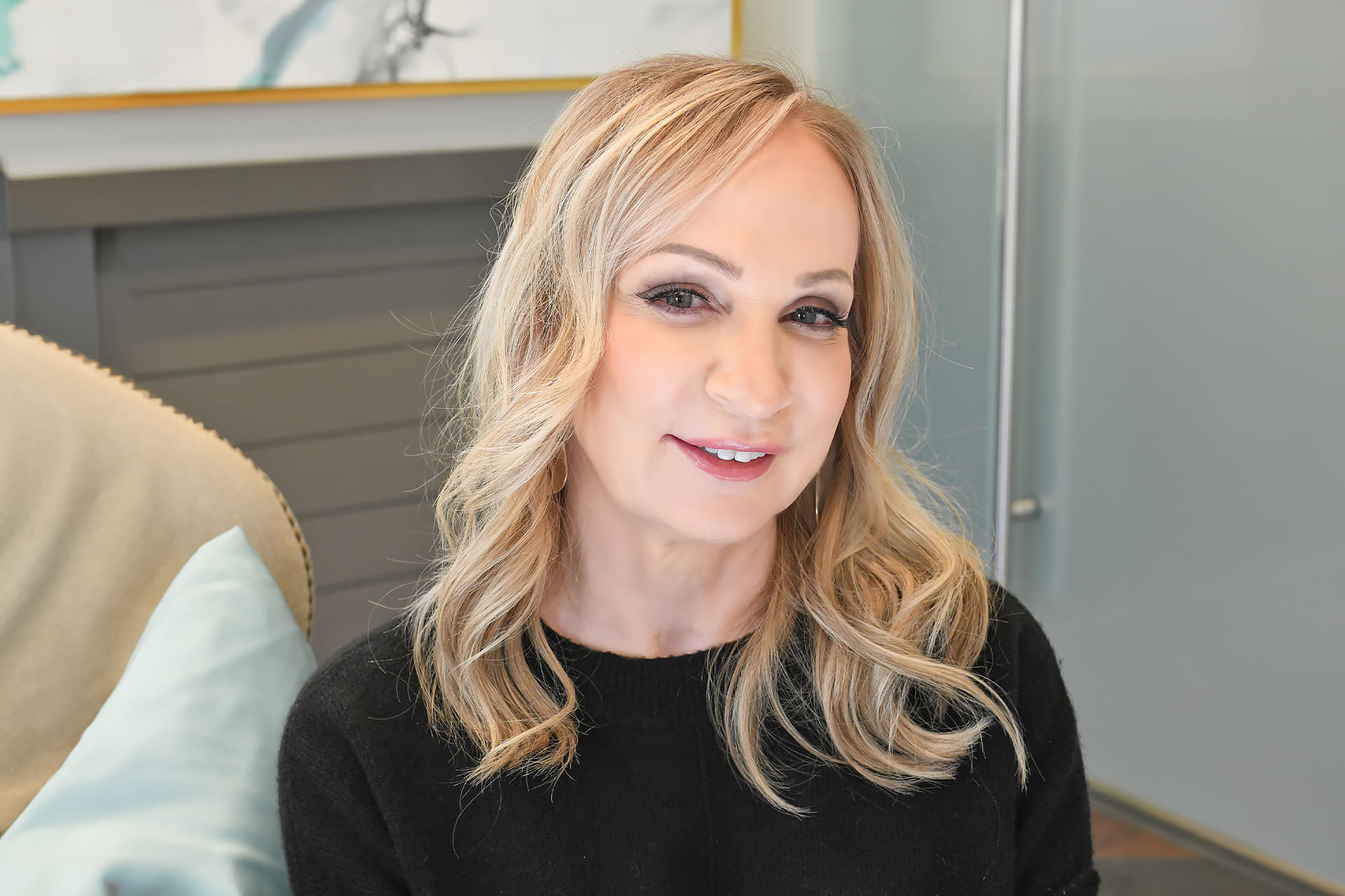 A woman with shoulder-length blonde hair is sitting indoors. She is wearing a black sweater and smiling slightly at the camera. The background appears to be a modern room with a grey wall and glass panel, along with a painting partially visible.