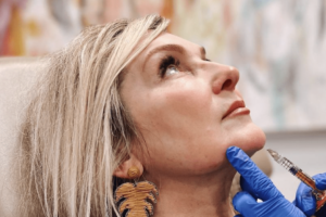 A person with light-colored hair is seated and looking upwards. Another person wearing blue gloves is seen holding a syringe near the seated person's chin. The background is blurred with various colors.