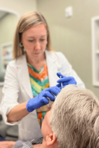 A healthcare professional wearing a white coat and blue gloves is administering an injection to an elderly person who is seated. The healthcare provider is focused on the task, while the patient is looking up. The setting appears to be a medical office or clinic.
