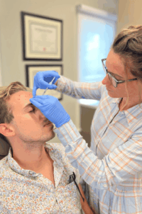 A person with short blonde hair and a patterned shirt is sitting in a chair, receiving an injection on the forehead from a healthcare professional wearing blue gloves, glasses, and a striped shirt. In the background, there are framed certificates on the wall and a window with a roller blind.
