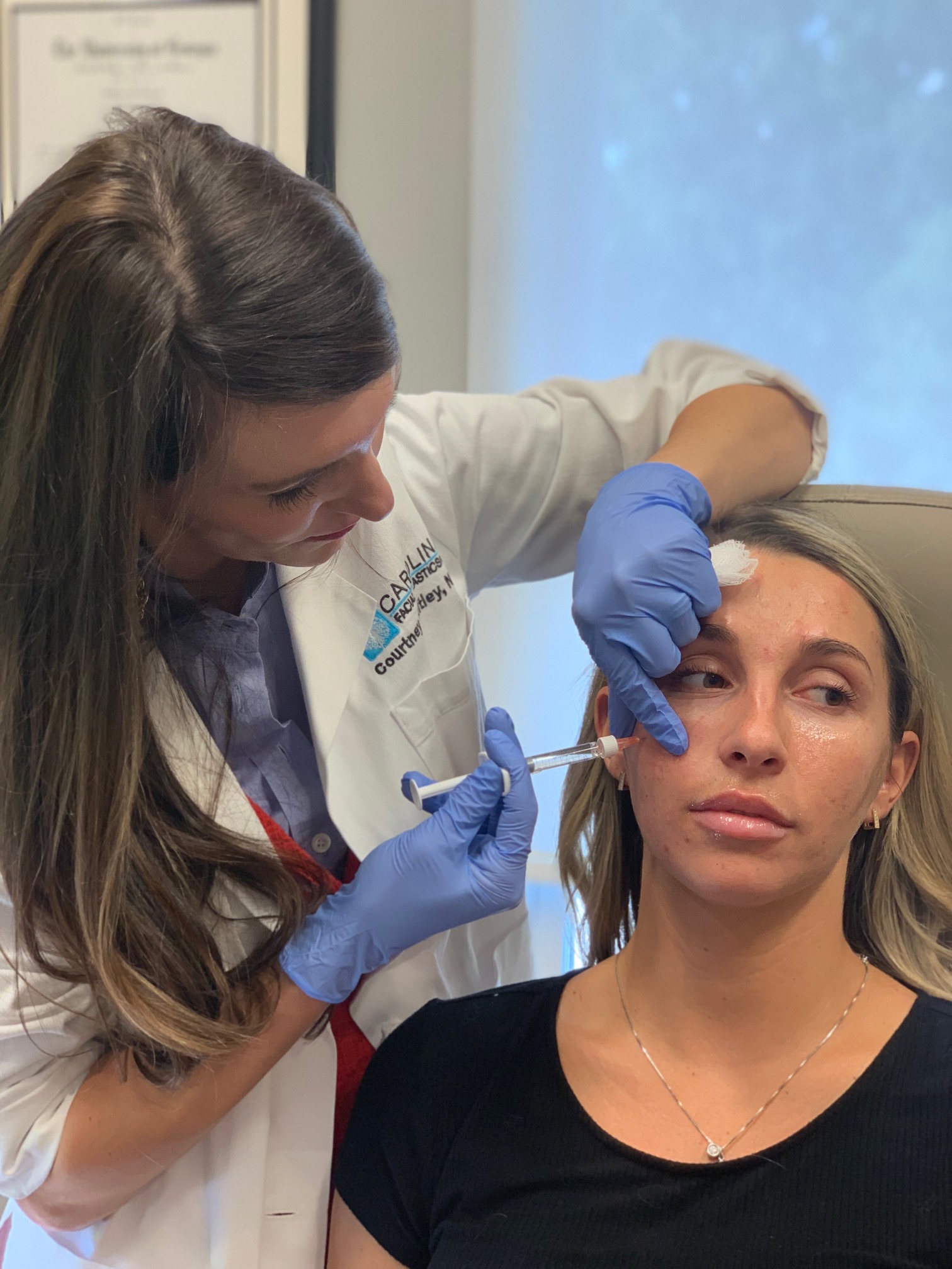 A woman in a medical office receiving a facial injection from a healthcare professional. The healthcare professional is wearing a white lab coat, gloves, and is applying the injection to the woman's cheek while holding gauze on her forehead. The woman receiving the injection looks relaxed and is sitting in a reclining chair.