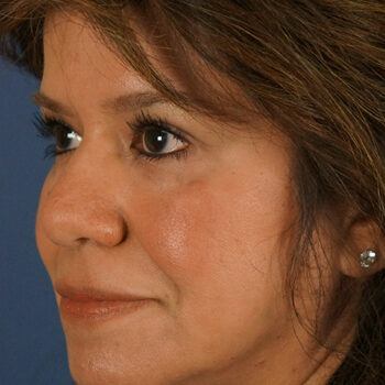 Close-up of a woman with shoulder-length brown hair and dark eyes, showing a side profile of her face. She is looking slightly to the left and has a subtle smile. The background is a plain blue color.