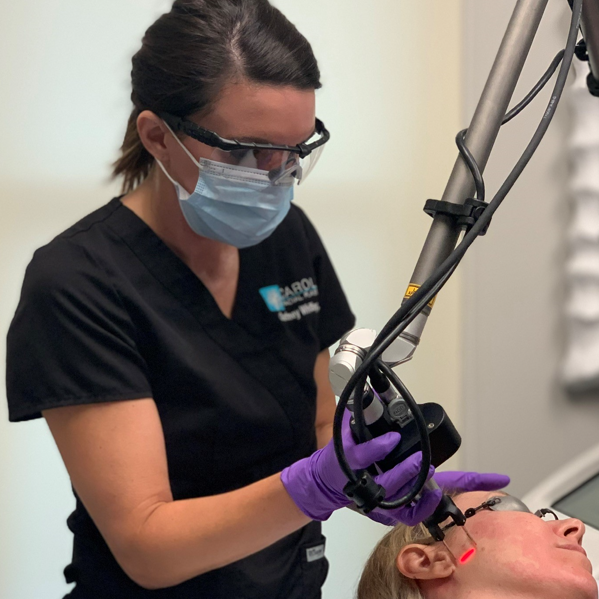 A medical professional wearing a mask, protective eyewear, and gloves uses a laser on a patient's face during a cosmetic procedure. The patient is lying down with their eyes protected while the procedure is being performed.