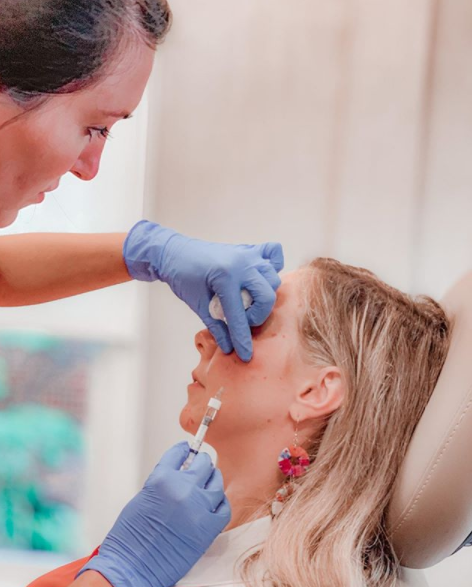 A person receives an injection in their face from a professional wearing blue gloves. The patient is seated and seems relaxed, with the professional focused on administering the injection. The setting appears to be a medical or cosmetic treatment room.