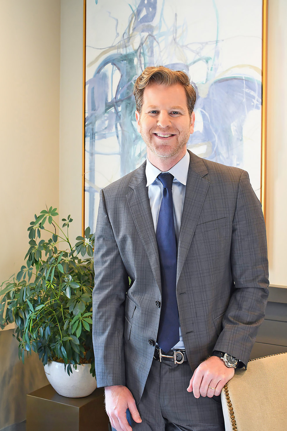 A man in a gray checked suit with a blue tie is standing and smiling in an indoor setting. Behind him, there is an abstract painting on the wall and a green potted plant on a table. He has short, styled hair and appears to be in a professional environment.