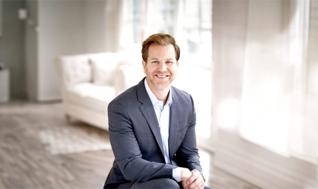 A smiling person wearing a dark suit with a white shirt is sitting on a chair in a brightly lit room. The room has large windows with white curtains, and a white sofa is visible in the background. The room has a modern and clean aesthetic with light-colored wooden flooring.
