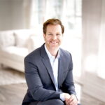 A smiling person wearing a dark suit with a white shirt is sitting on a chair in a brightly lit room. The room has large windows with white curtains, and a white sofa is visible in the background. The room has a modern and clean aesthetic with light-colored wooden flooring.