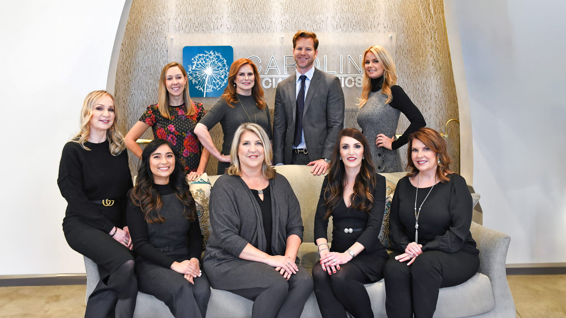 A group of nine individuals, consisting of eight women and one man, are posed in a well-lit office setting. Five women are seated on a couch, while three women and the man stand behind. The group is dressed in professional attire, standing in front of a wall with a sign that reads "Carolina Orthodontics." Everyone is smiling and looking towards the camera, conveying a friendly and welcoming atmosphere.